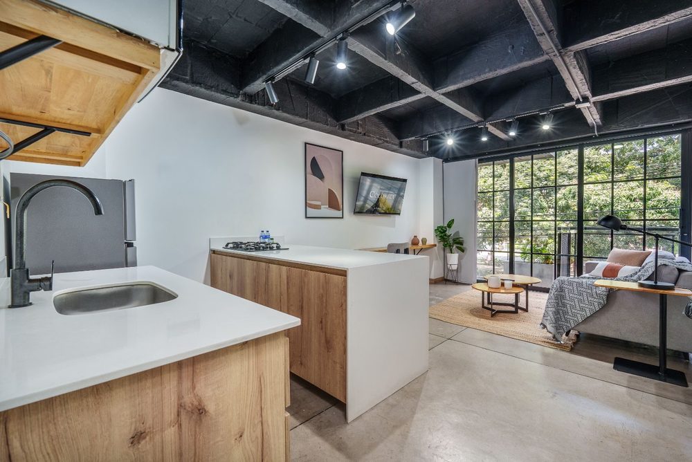 Cocina abierta con isla blanca, losa negra a la vista y sala junto a un ventanal de piso a techo